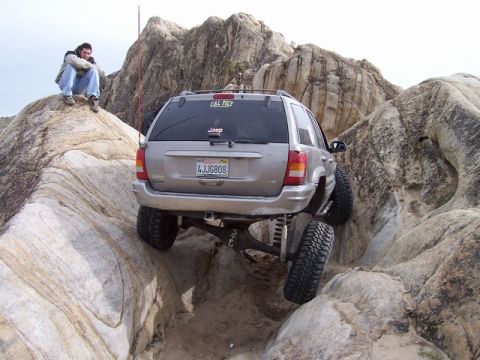 1989 Ford Bronco - A silver Jeep Grand Cherokee is shown off-roading through a rocky landscape, with a person sitting on a boulder nearby.