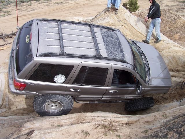Jeep Grand Cherokee on rocks, one wheel off the ground. Two people watching
