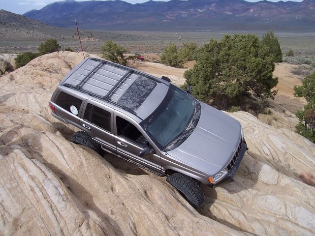 Silver Jeep Grand Cherokee off-roading on a rocky incline, with a roof rack and aftermarket tires