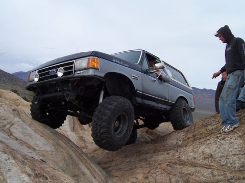 1989 Ford Bronco - This lifted Ford Bronco is navigating a rocky off-road trail with people watching.