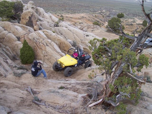 Yellow off-road buggy with roll cage on a rocky hill; person sits on ground taking photos. Silver SUV visible in background