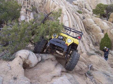 1989 Ford Bronco - A yellow off-road vehicle navigates a rocky, desert terrain with a person watching in the background.
