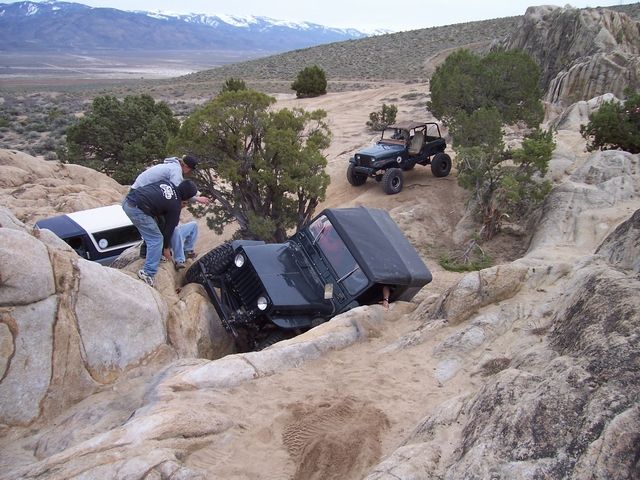 Dark green Jeep CJ-7 tilted in a rocky ditch with a person inside, another Jeep behind it, and a white and blue vehicle partially visible