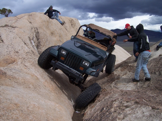 Teal Jeep Wrangler YJ crawling nose-down through granite rock crevice with spotters watching