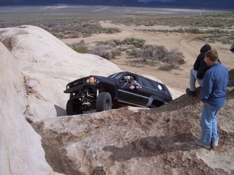 1989 Ford Bronco - A black SUV is shown off-roading through a rocky landscape, with two people observing.