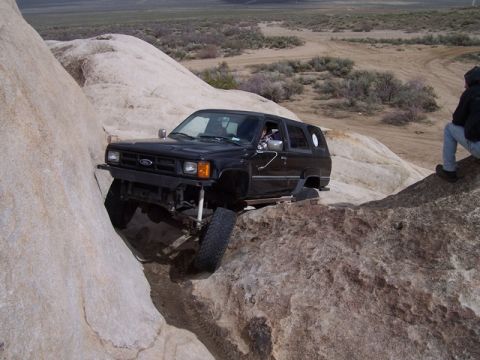 1989 Ford Bronco - A black SUV is shown attempting to maneuver through a challenging rocky terrain, with a person observing from a nearby rock.