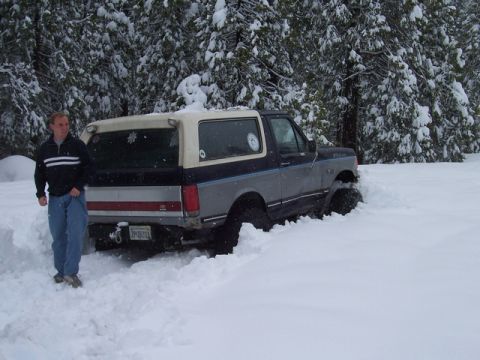 1989 Ford Bronco - A man stands beside a 1989 Ford Bronco that is stuck in deep snow, with trees covered in snow in the background.
