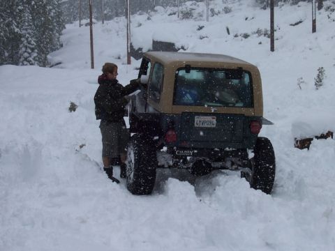 1989 Ford Bronco - A person attempts to get into a Jeep Wrangler that is partially buried in snow.