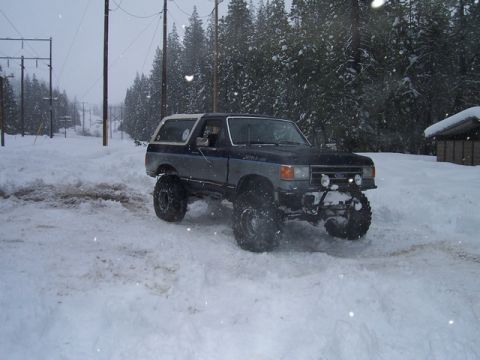 1989 Ford Bronco - A lifted Ford Bronco navigates a snowy road with trees and power lines in the background.