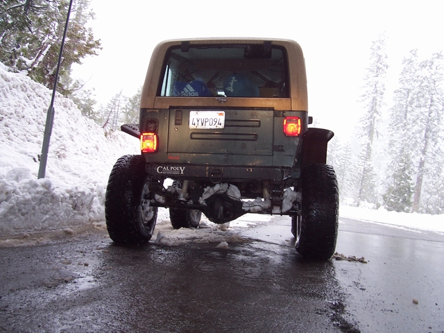 Olive green Jeep Wrangler with a tan hardtop, lifted suspension, and large off-road tires, seen from the rear in a snowy environment