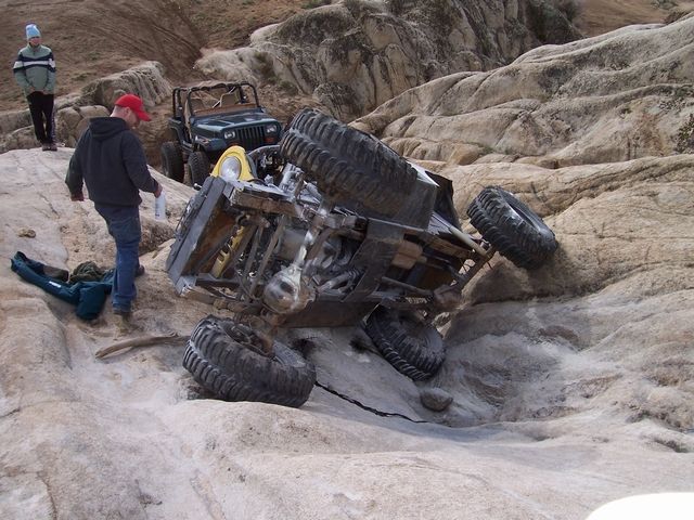 Off-road vehicle tipped over on rocks with another Jeep nearby. People are standing around observing
