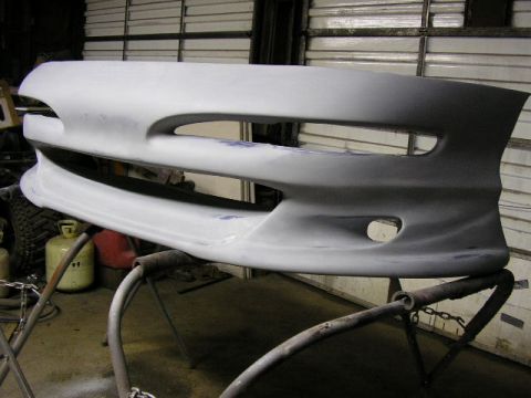1994 Ford Probe - A gray, unpainted front bumper cover is shown mounted on a stand in a workshop.