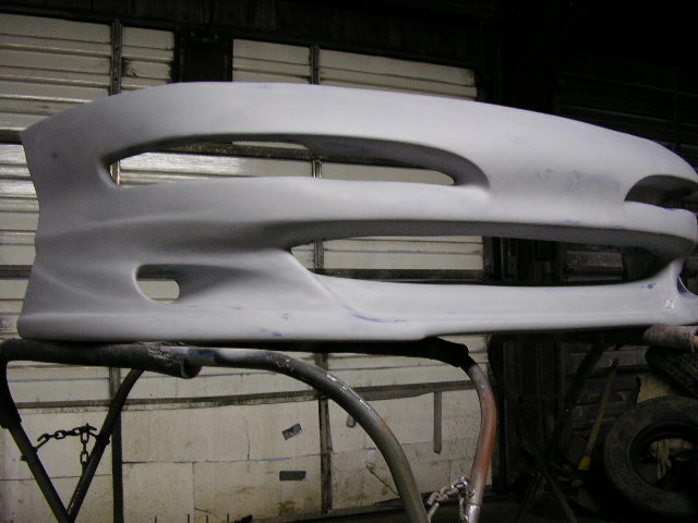 Unpainted car bumper on a stand, showing large central grille opening and two smaller side openings