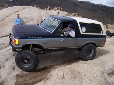 1989 Ford Bronco - A modified Ford Bronco is driven off-road with a person inside and another standing nearby.