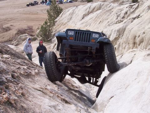 1989 Ford Bronco - Jeep Wrangler navigates a challenging rocky terrain with spectators observing the background shows multiple vehicles and tents.
