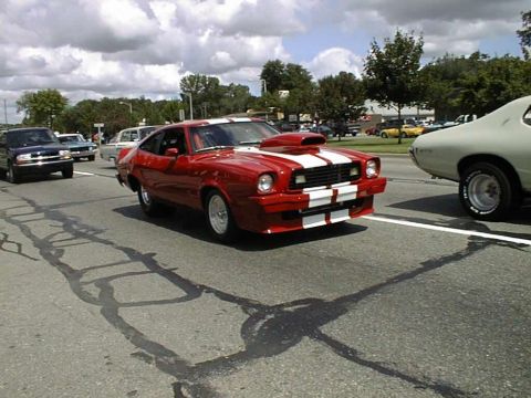 1978 Ford Mustang II - A red coupe with racing stripes and a prominent hood scoop drives down the road.