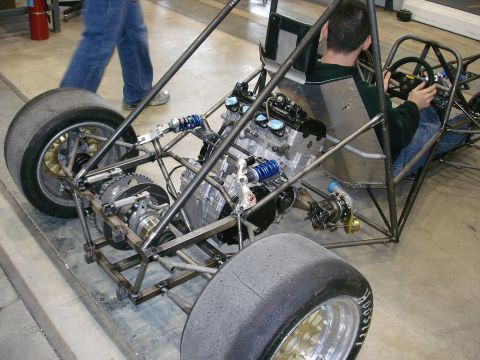 1985 Ford LTD Crown Victoria - A driver sits in the cockpit of a custom-built open-wheel race car chassis, its engine and suspension components.