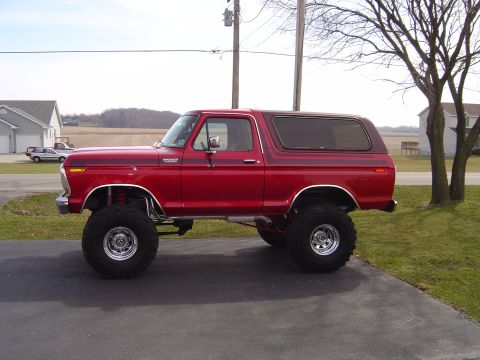 1978 Ford Bronco - Full side profile of a lifted red full-size Ford Bronco with its hardtop on, parked on an asphalt driveway with bare trees and farm fields…