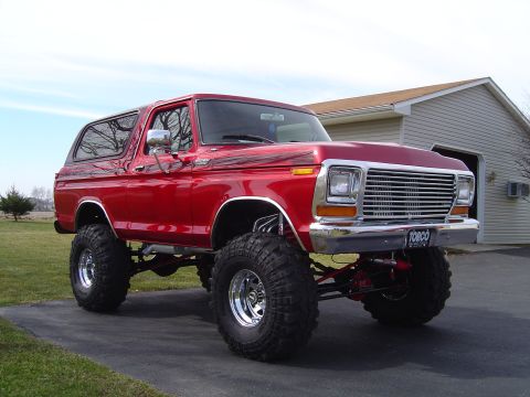 1978 Ford Bronco - Three-quarter front view of a lifted red full-size Ford Bronco with hardtop, parked on a driveway in front of an open garage. It wears a ch…