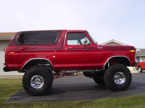 1978 Ford Bronco - Passenger-side profile of a lifted candy-red full-size Ford Bronco with its hardtop installed, parked on a driveway between houses. Chrome…