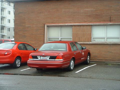 1999 Ford Crown Victoria - A red sedan is parked next to another red car in front of a brick building.