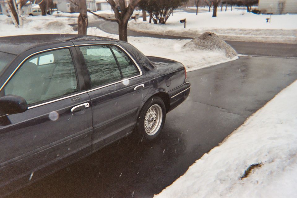 Dark blue Ford sedan on wire-spoke wheels parked on a wet snowy driveway during light snowfall