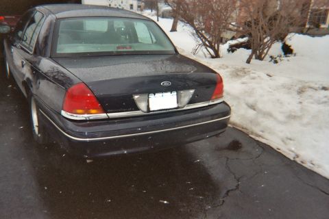 2000 Ford Crown Victoria - The rear of a dark Ford Crown Victoria is visible, parked on a wet driveway with snowbanks nearby.