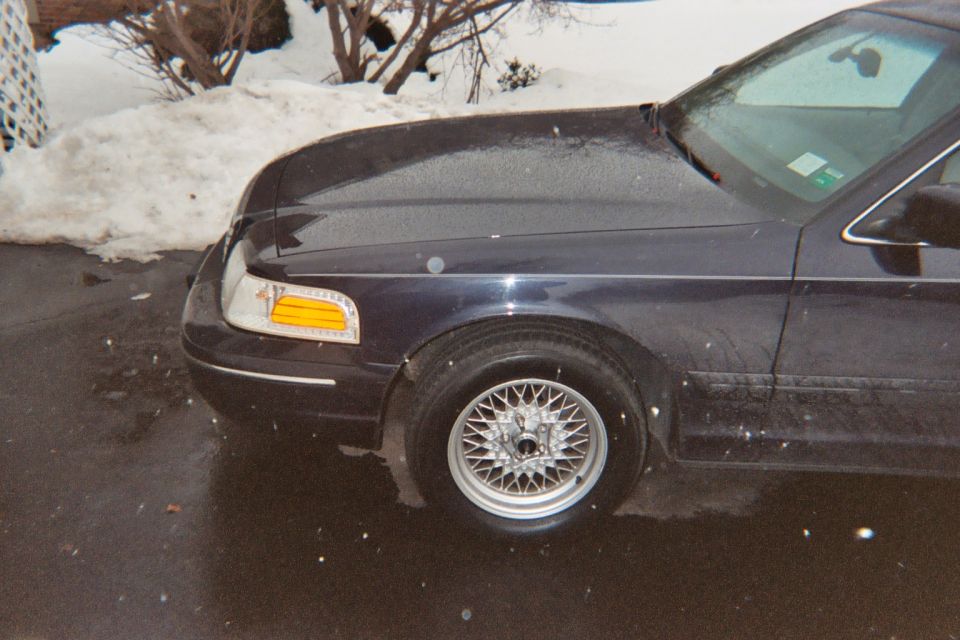 Front corner of a dark Ford Crown Victoria with wire wheel cover on a snowy driveway