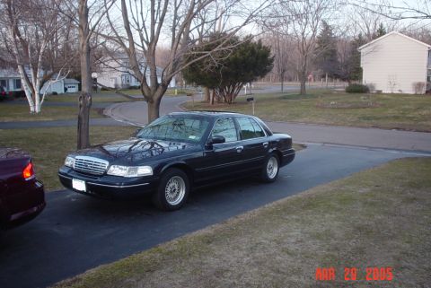 2000 Ford Crown Victoria - A dark blue/black Ford Crown Victoria full-size sedan parked in a residential driveway, shown from a front three-quarter angle with chrome…