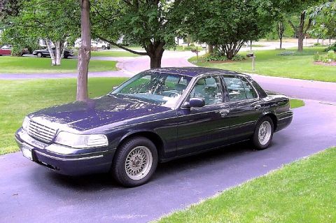 2000 Ford Crown Victoria - A dark blue Ford Crown Victoria full-size sedan parked on a paved driveway, shown from a front three-quarter angle with wire-spoke wheels a…