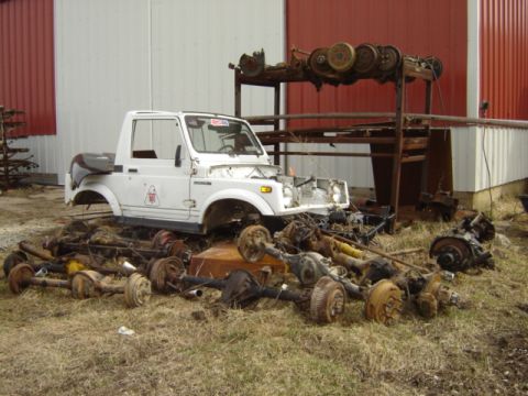 1992 Ford Bronco - A white Suzuki Samurai sits in a yard piled high with various axles and vehicle parts.