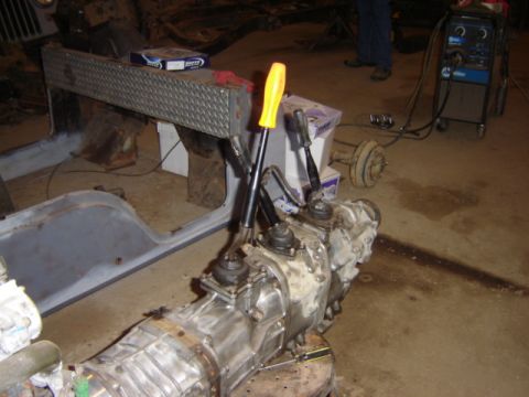 1992 Ford Bronco - Transmission and transfer case assembly with shifters, next to a disassembled vehicle body in a workshop.