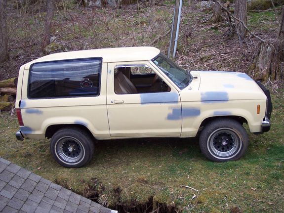Side view of cream Ford Bronco II SUV with gray primer spots and black steel wheels parked on grass