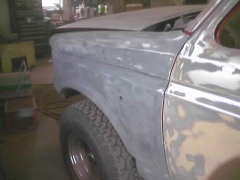 1989 Ford Bronco - Bodywork in progress on a truck's front fender and door, shot from the front corner inside a shop: the panels are sanded down to gray prime…