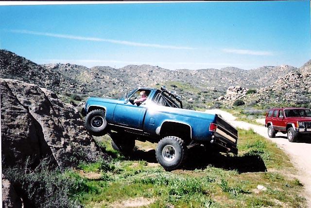 Blue lifted Chevrolet pickup climbing rocks off-road with a red Jeep behind on a dirt trail