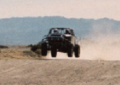 1990 Ford Bronco - dark off-road race vehicle launches over a desert rise, throwing up a cloud of dust against distant brown hills.