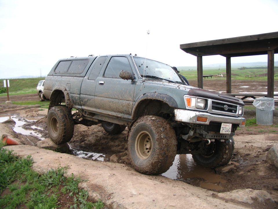 Lifted green Toyota pickup with camper shell stopped in muddy ruts near open shelter