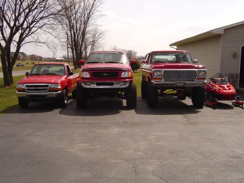 1978 Ford Bronco - Three red Ford trucks lined up front-on in an asphalt driveway: a stock Ranger, a lifted F-150 with a black hood bra and oversized tires, a…