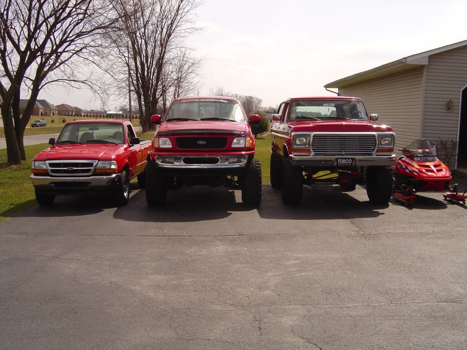 Three red Ford trucks parked front-on in a driveway with a red snowmobile at right