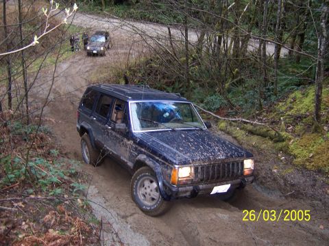 1987 Jeep Cherokee - A dark blue 1987 Jeep Cherokee navigates a muddy off-road trail with another vehicle visible in the background.