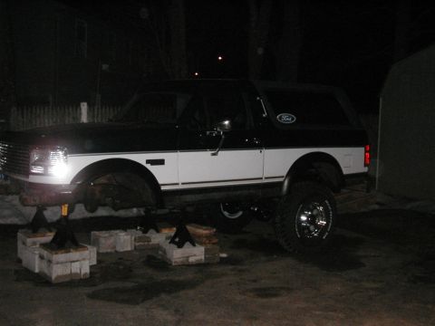 1994 Ford Bronco - A two-tone 1994 Ford Bronco is raised on jack stands in a dimly lit outdoor area, suggesting maintenance or modification work.