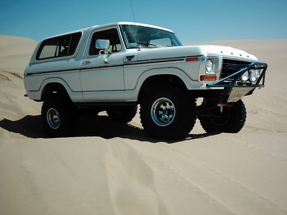 White 1978 Ford Bronco with off-road tires and a brush guard, parked on sand dunes
