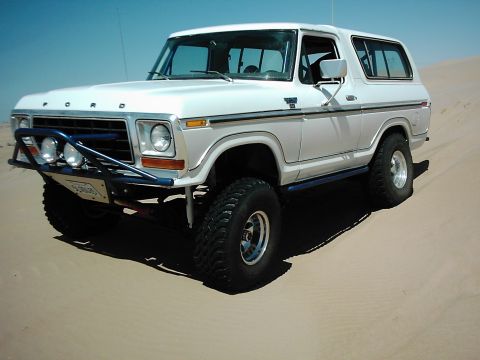 1978 Ford Bronco - This white 1978 Ford Bronco features a custom blue bumper and off-road tires, seen here on a sandy terrain.