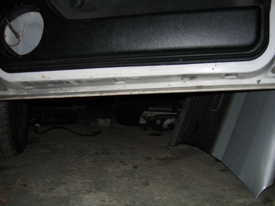 Open vehicle door over white rocker panel, exhaust and undercarriage visible beneath in dim garage