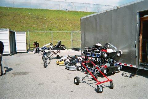 1990 Ford Bronco - Several racing go-karts and a BMX bike sit on stands in a paved lot beside a gray enclosed trailer and a grassy embankment.