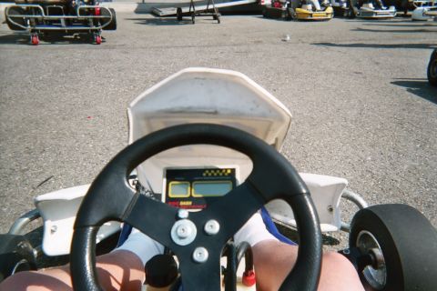 1990 Ford Bronco - View from a go-kart driver's seat over the black steering wheel and small digital dash, with other karts parked on the pavement ahead.
