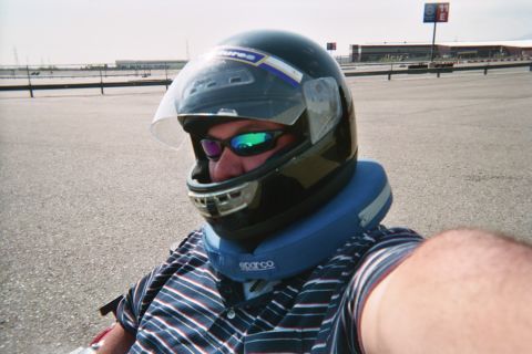 1990 Ford Bronco - man wearing a dark full-face racing helmet, mirrored sunglasses and a blue neck collar takes a selfie on an open paved track.