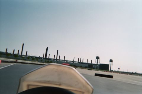 1990 Ford Bronco - Forward view over a go-kart's nose down an open paved track toward distant grandstands, light poles and water towers under a clear sky.