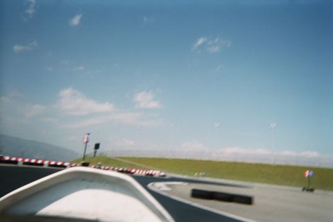 1990 Ford Bronco - Motion-blurred view over a go-kart nose along a curving track lined with red-and-white barriers, with mountains on the horizon.
