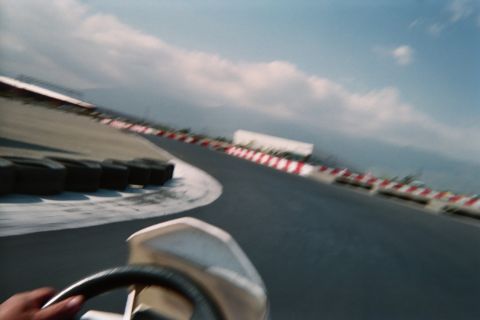 1990 Ford Bronco - Motion-blurred cockpit view of a go-kart rounding a curve past tire stacks and red-and-white barriers under a cloudy sky.